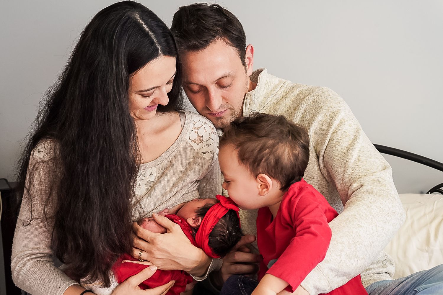 Young family with parents and toddler cuddling newborn baby indoors in warm embrace.