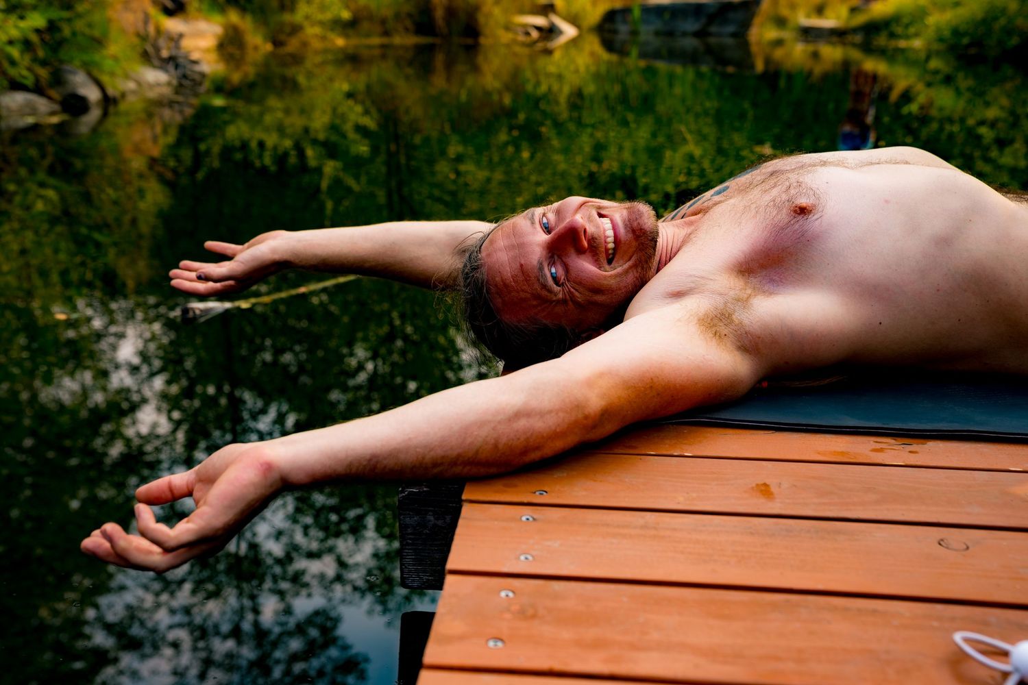 Shirtless man lying on a wooden dock by a lake, arms outstretched, relaxing outdoors in nature.