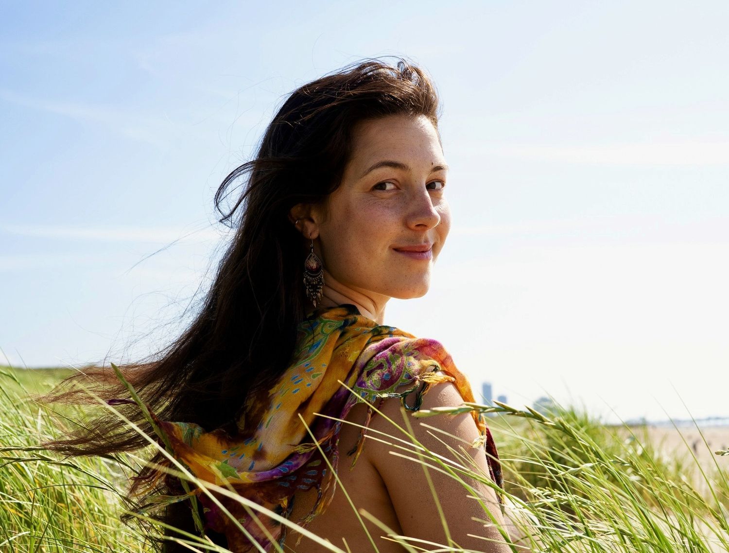A photo of a woman with brown hair, a colorful scarf, grass, blue skies, and gracefulness with the wind blowing.
