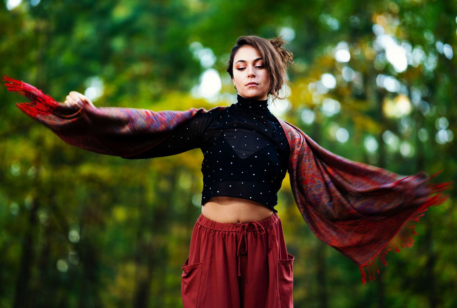 A photo of a woman wearing a black top with red pants and a scarf is draped around her and she is looking down.