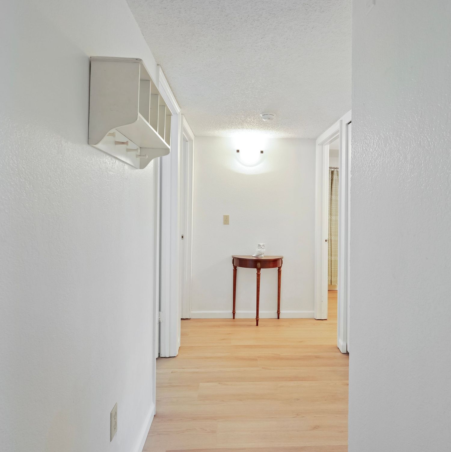 Bright hallway with hardwood floors, white walls, a small red table at the end, and a medicine cabinet on the left wall.