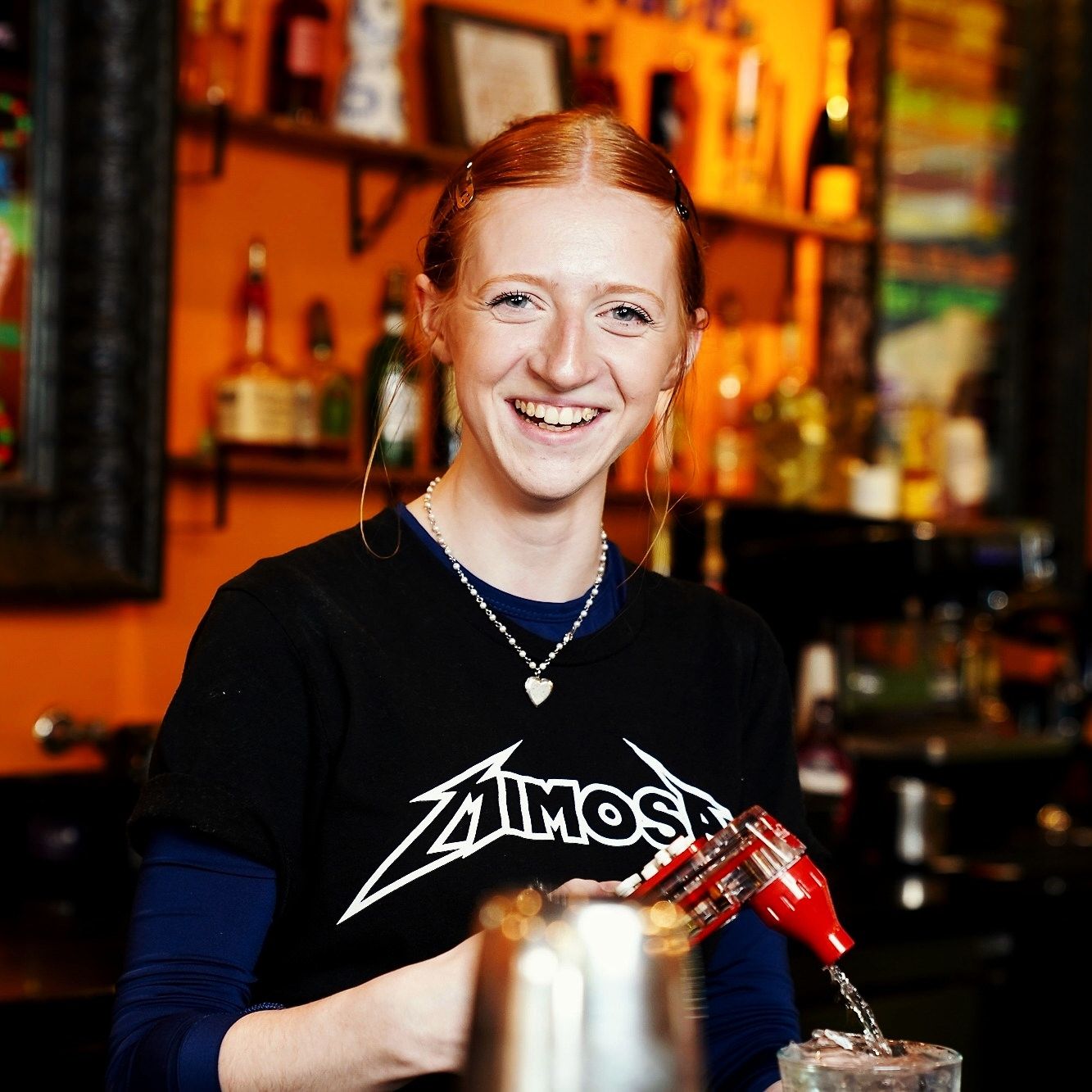 Smiling female bartender in black Mimosa shirt pouring a cocktail at a vibrant bar.