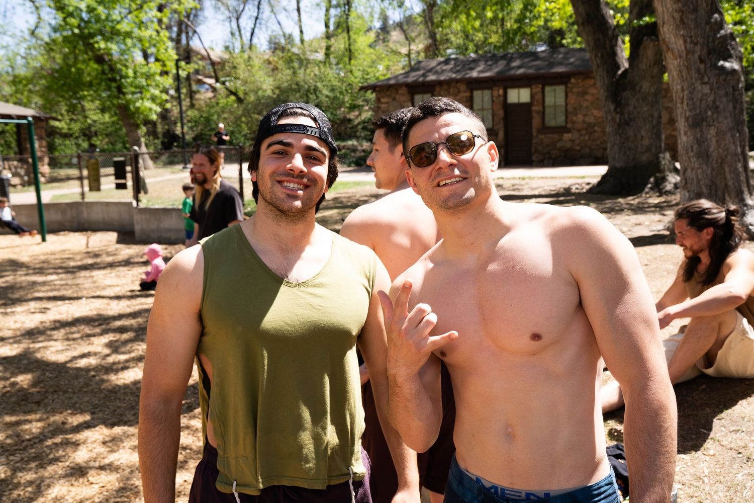 Two shirtless men smiling outdoors at a sunny park gathering with trees and people in the background.