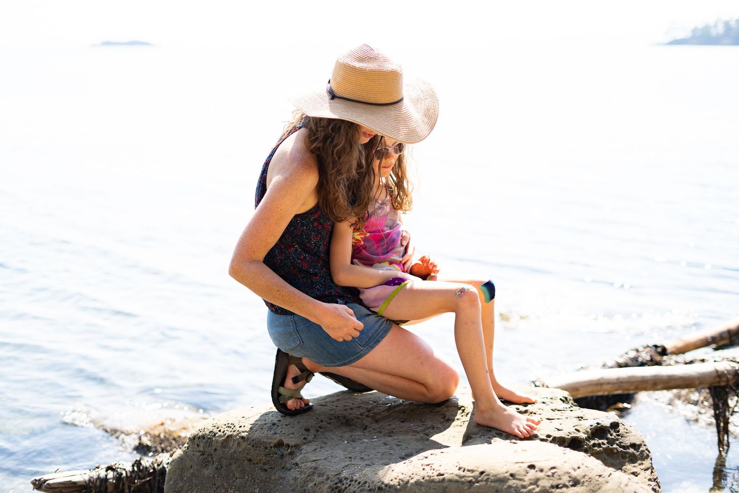A mother in a straw hat and denim shorts sits on rocks by the ocean with her young child on a sunny day.