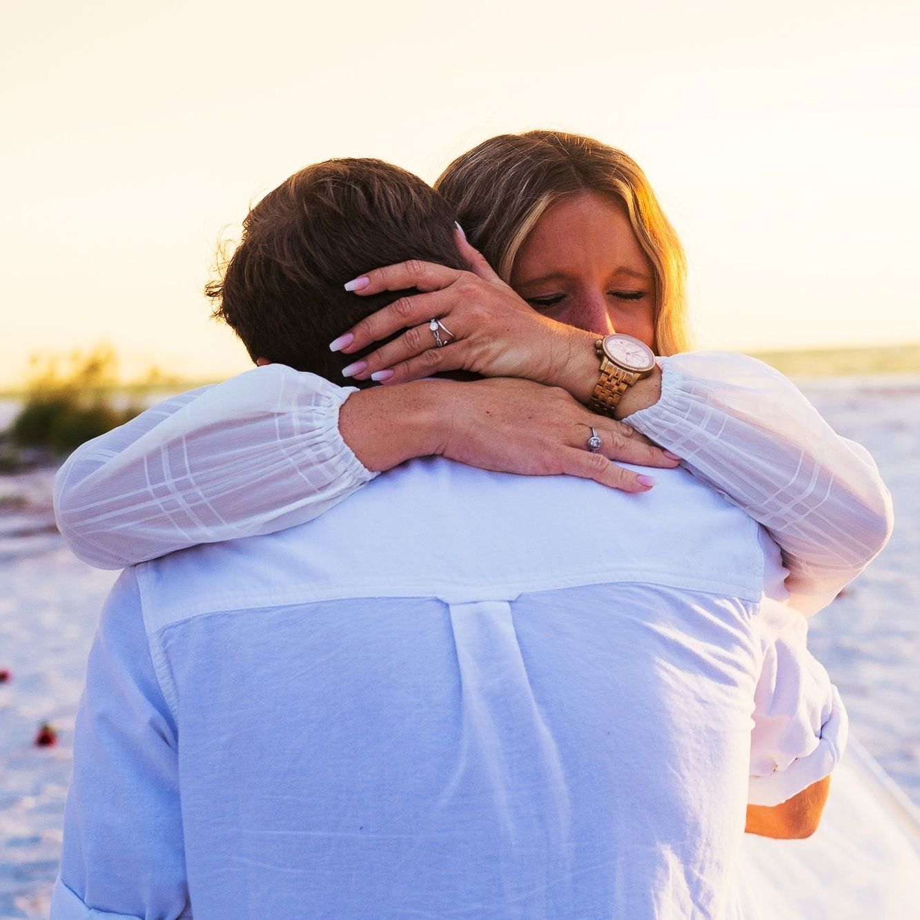 Couple sharing a tender beach hug at sunset, woman wearing a watch and rings, both dressed in light blue and white.