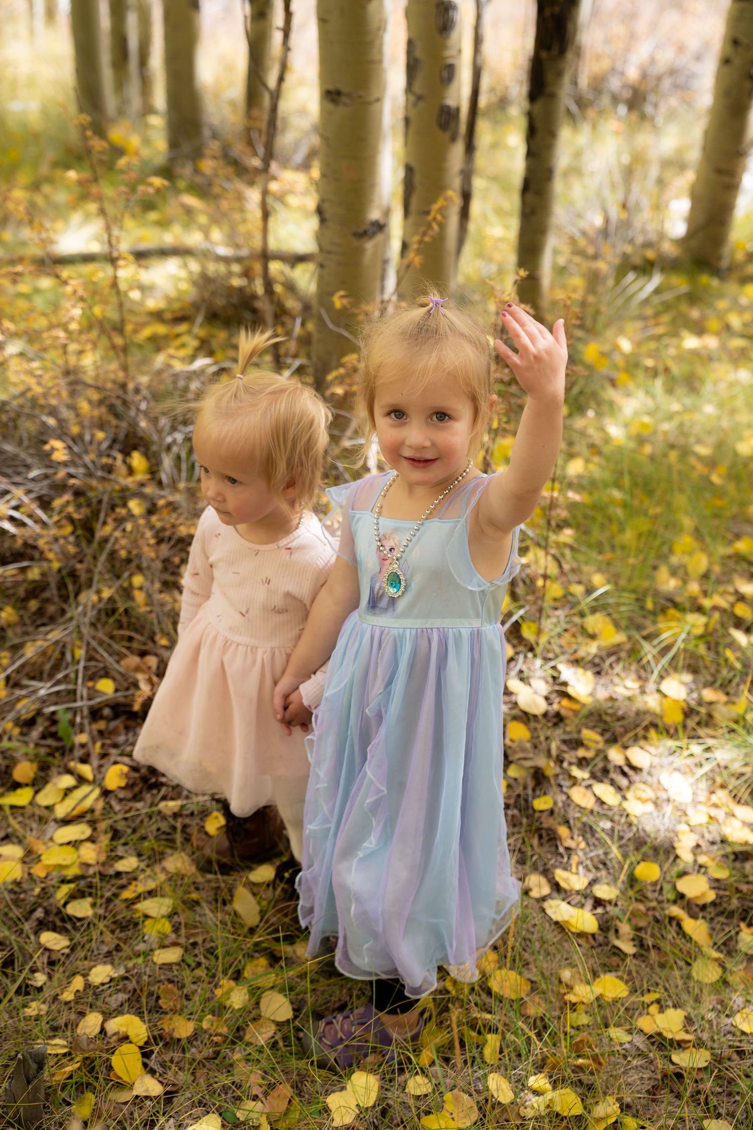 Two young children in light dresses play together among yellow fallen leaves in an autumn forest setting.