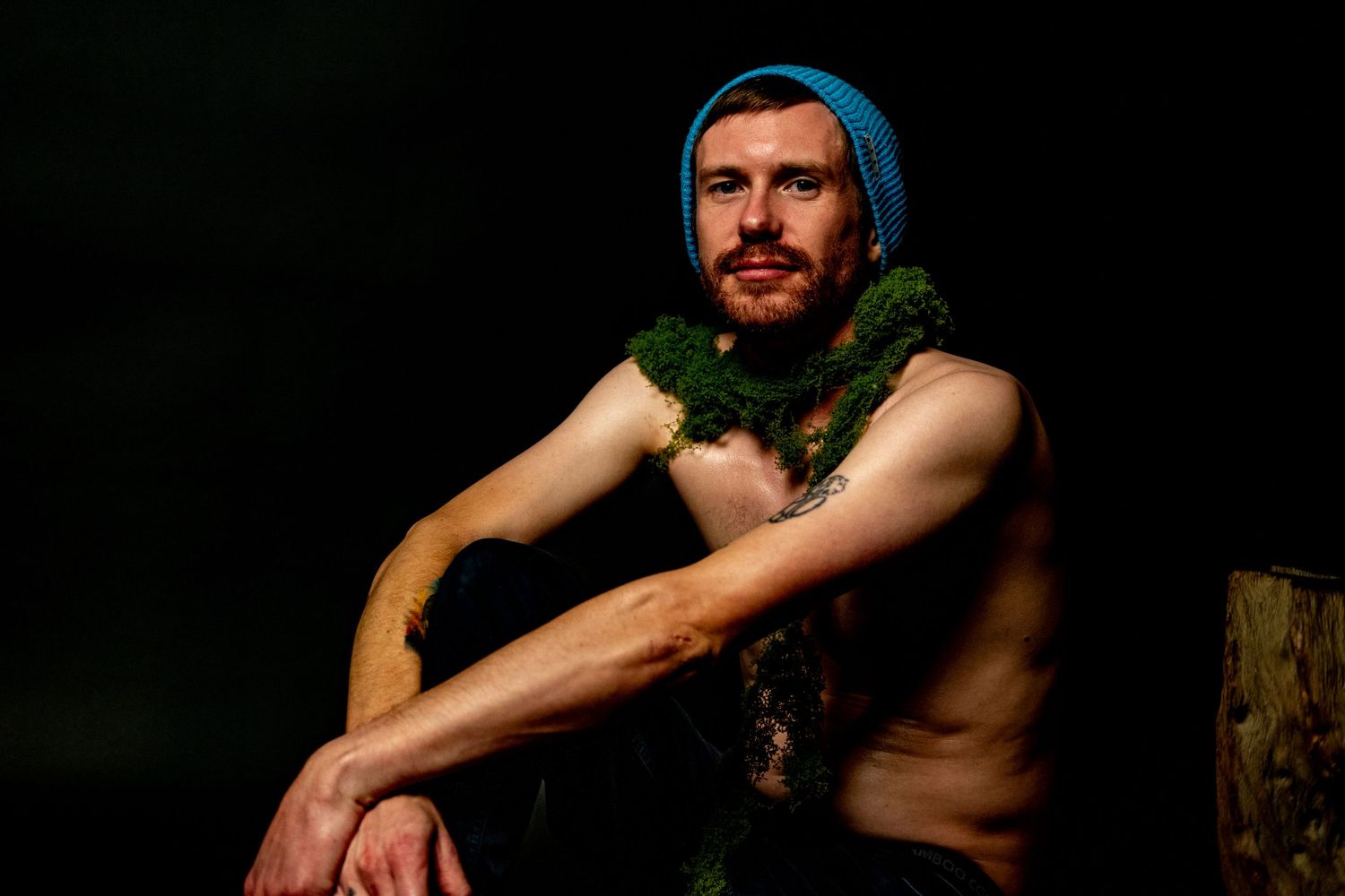A shirtless person wearing a blue beanie and green lei sits against a dark background in dramatic lighting.