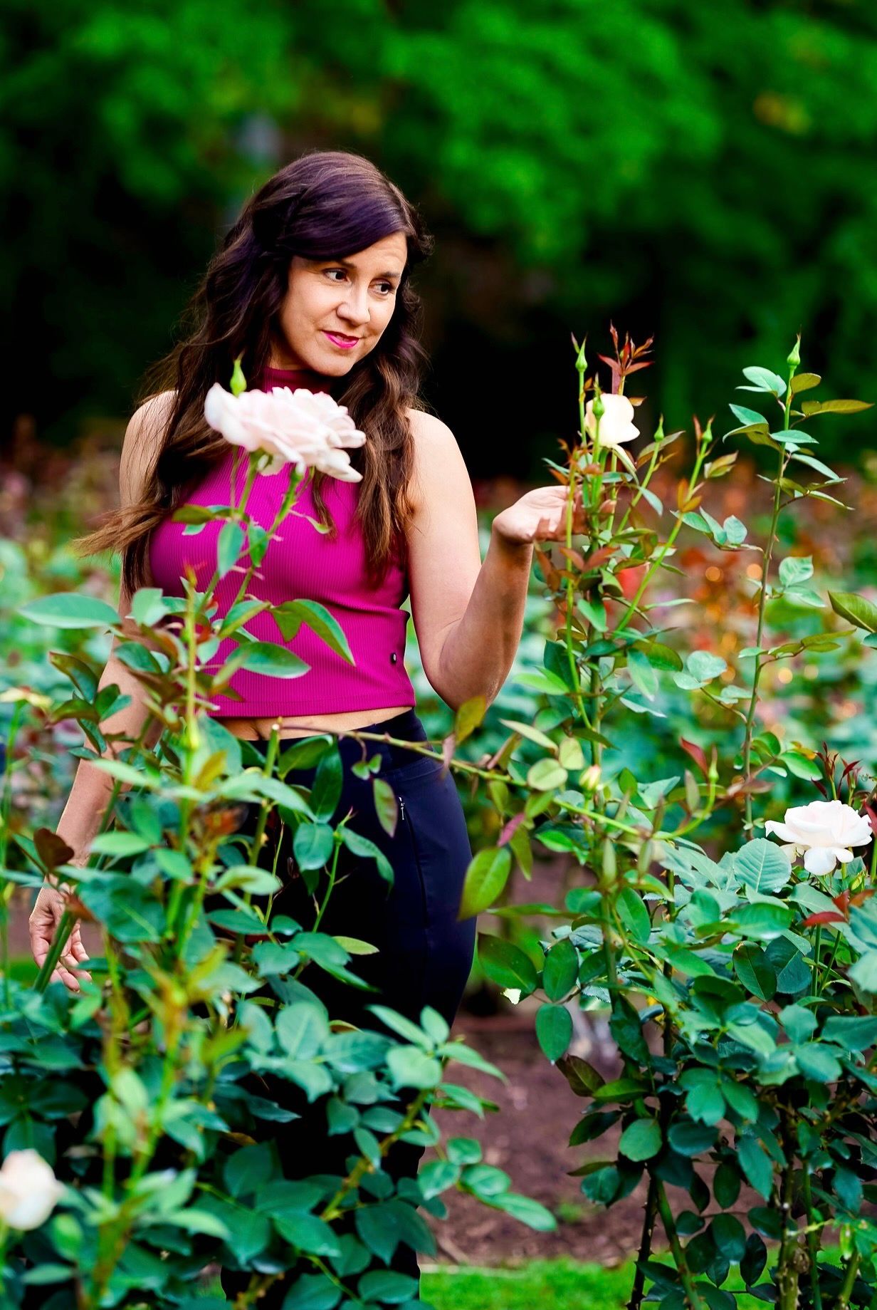 A woman in a magenta top poses among lush green foliage and white roses in a garden setting.