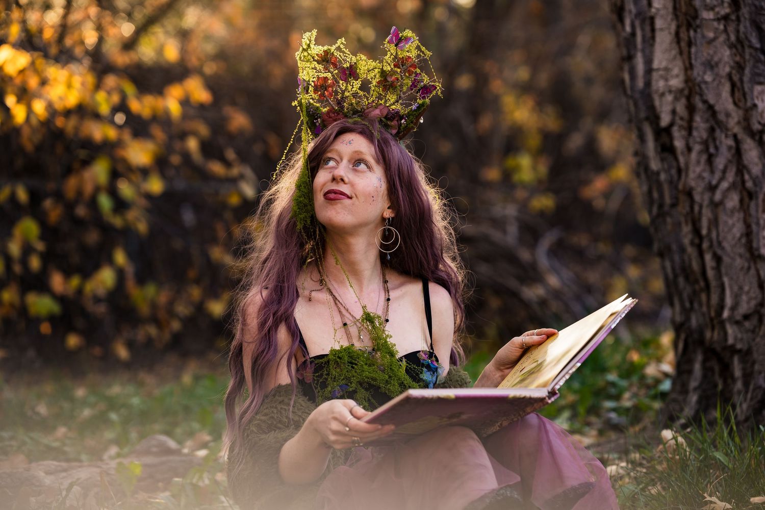 Woman in fantasy fairy costume with floral crown reads a book outdoors in an autumn forest setting.