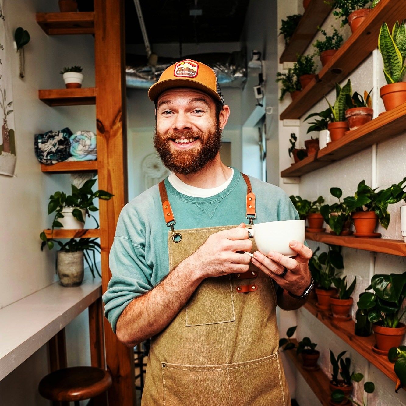 Bearded barista in apron and cap holding a coffee cup in a plant-filled cafe.