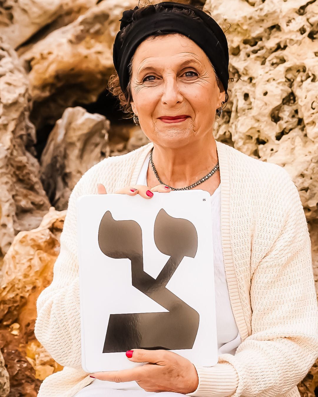 An older woman holds a Hebrew letter sign while sitting against a rocky background outdoors.