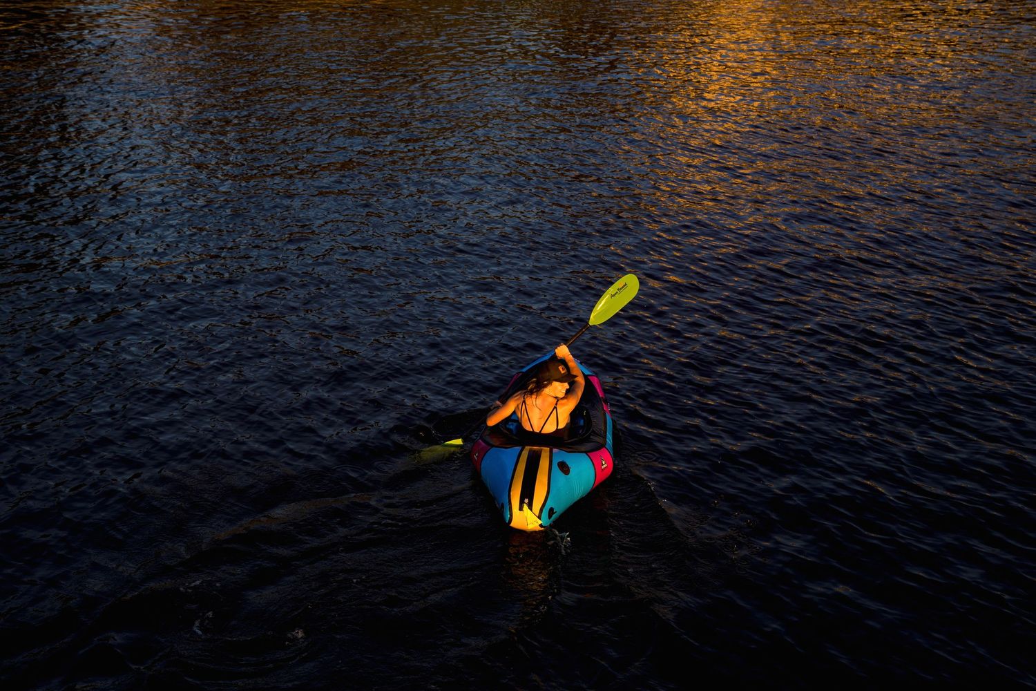 Person kayaking on dark water at sunset, paddling a colorful kayak with a yellow oar in golden light.