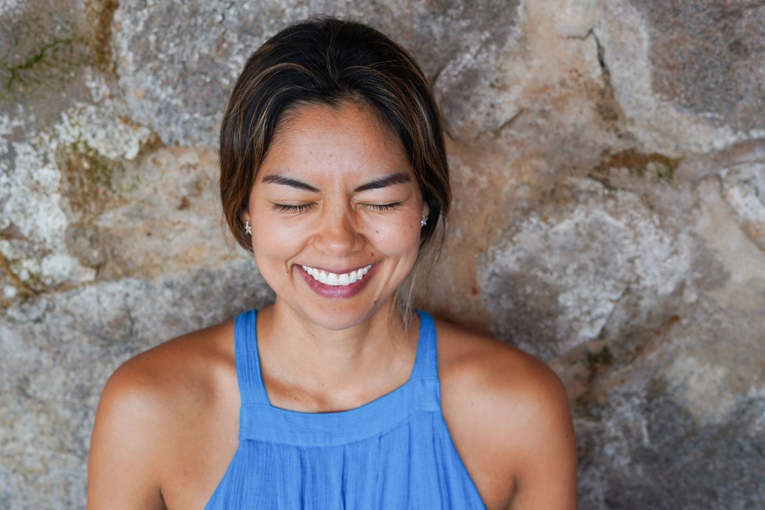 Young woman laughing joyfully with eyes closed, wearing a blue tank top against a rustic stone wall background.