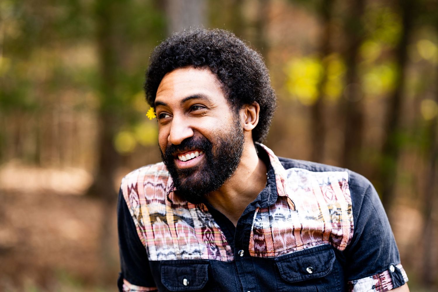 Smiling bearded Black man with afro laughing outdoors in a forest setting, wearing a colorful patterned shirt.