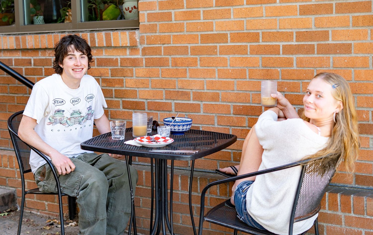 Two young people sitting at an outdoor patio table against a brick wall, smiling and enjoying drinks together.