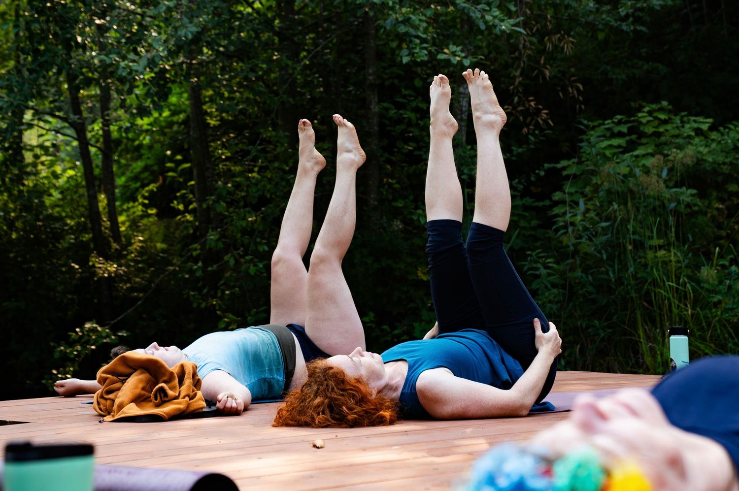 Two women practicing yoga outdoors on a wooden deck, legs raised in the air, surrounded by lush green trees.