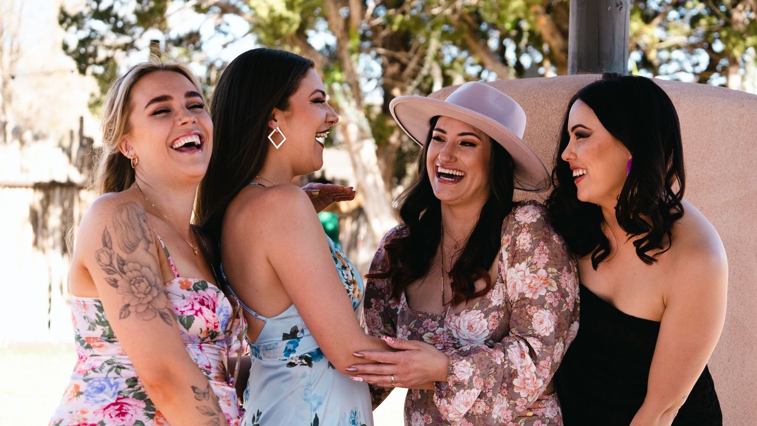 Four women laughing and embracing outdoors in floral and formal dresses at a sunny celebration event.