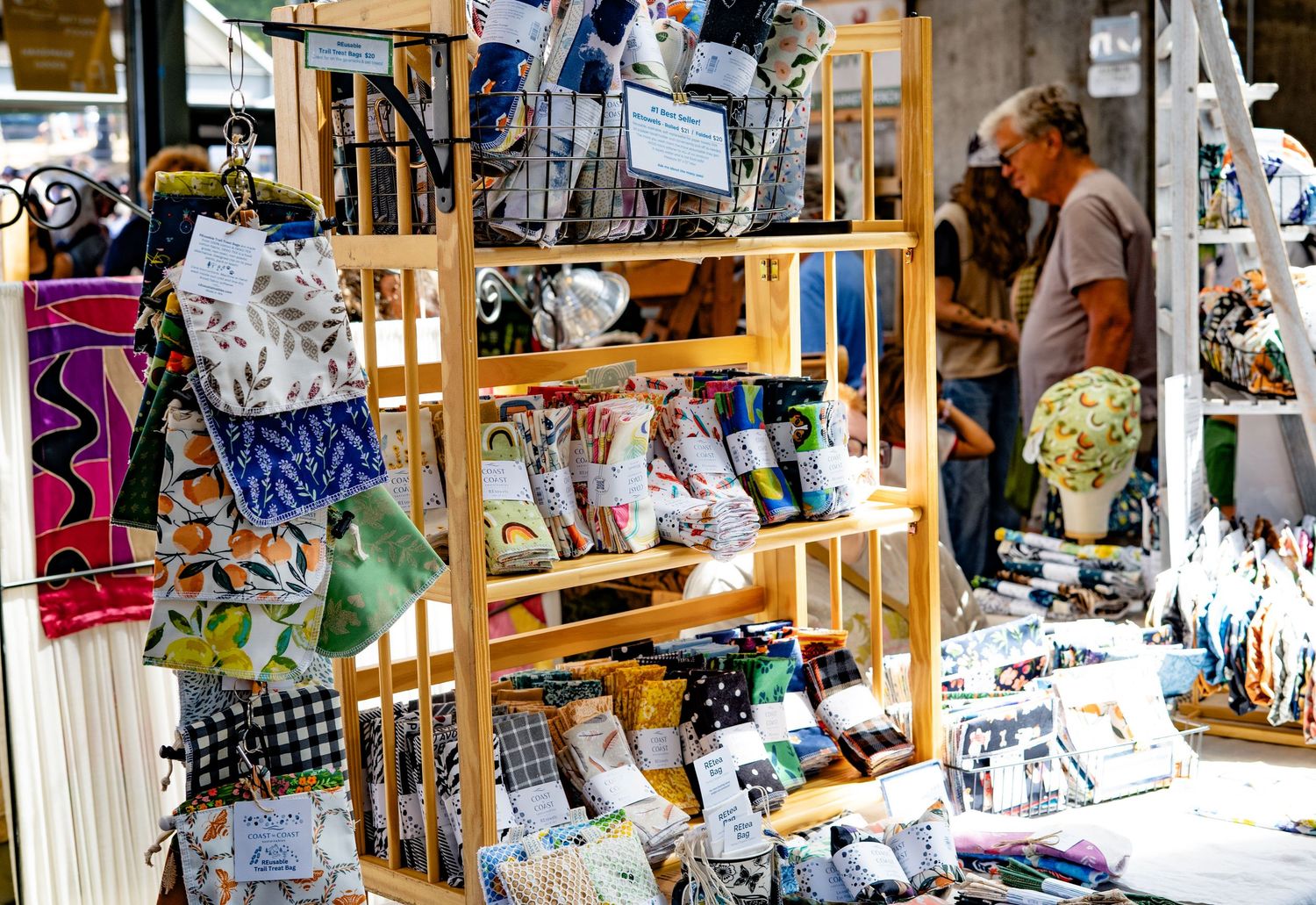 Wooden display shelves filled with handmade fabric goods at an outdoor craft market with shoppers browsing in background.