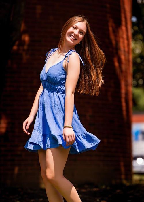 Young woman smiling in a blue sundress, posing outdoors near a brick wall on a sunny day.