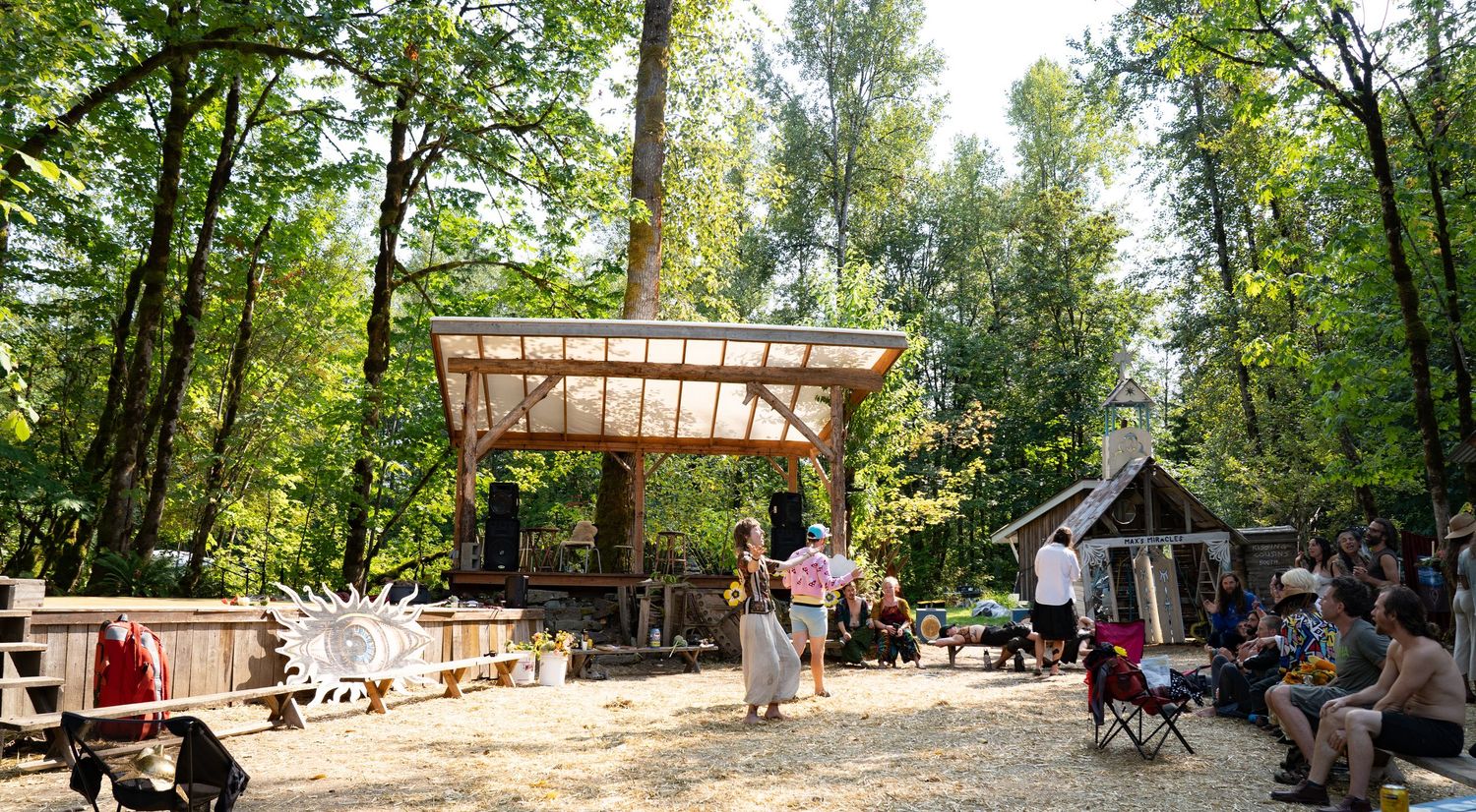 Outdoor woodland gathering with people relaxing near a rustic open-air pavilion surrounded by lush green trees on a sunny day.