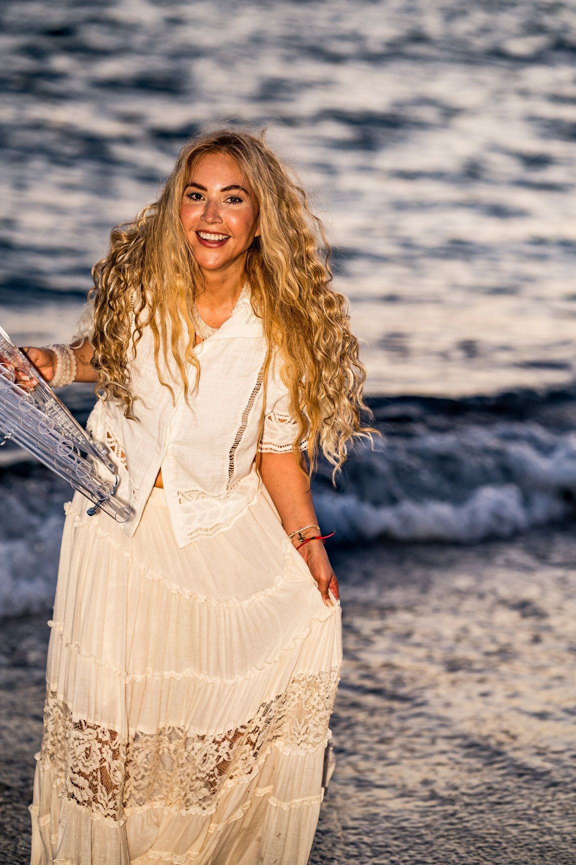 A photo of a woman wearing a white outfit, she has flowy blonde hair and has the ocean waves behind her.