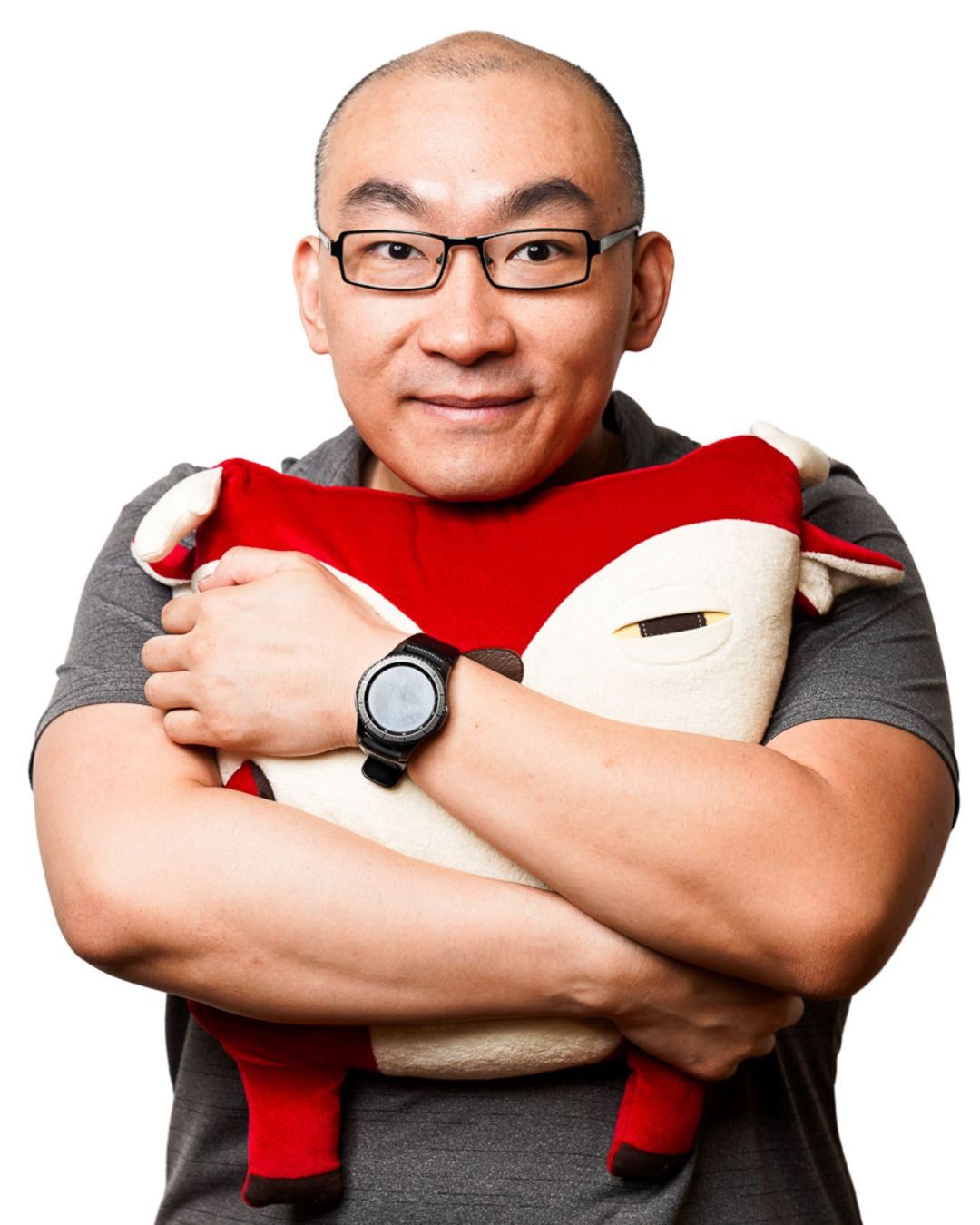 Smiling bald man with glasses hugging a red and white fox stuffed animal toy, wearing a gray shirt and watch.