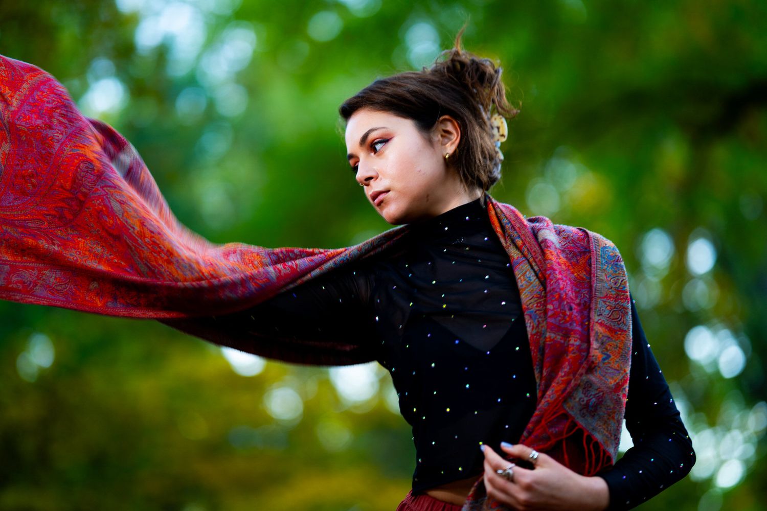 Young woman in black top with vibrant red flowing scarf outdoors among green trees.