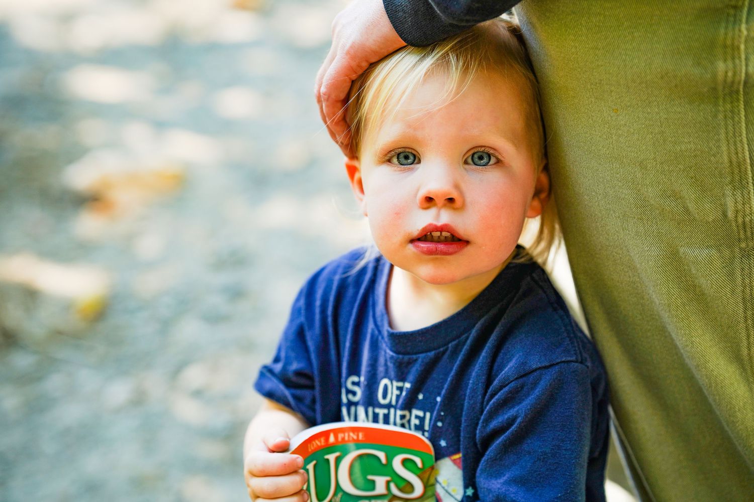 Toddler with blue eyes in a navy USGS shirt clinging to an adult's leg outdoors.