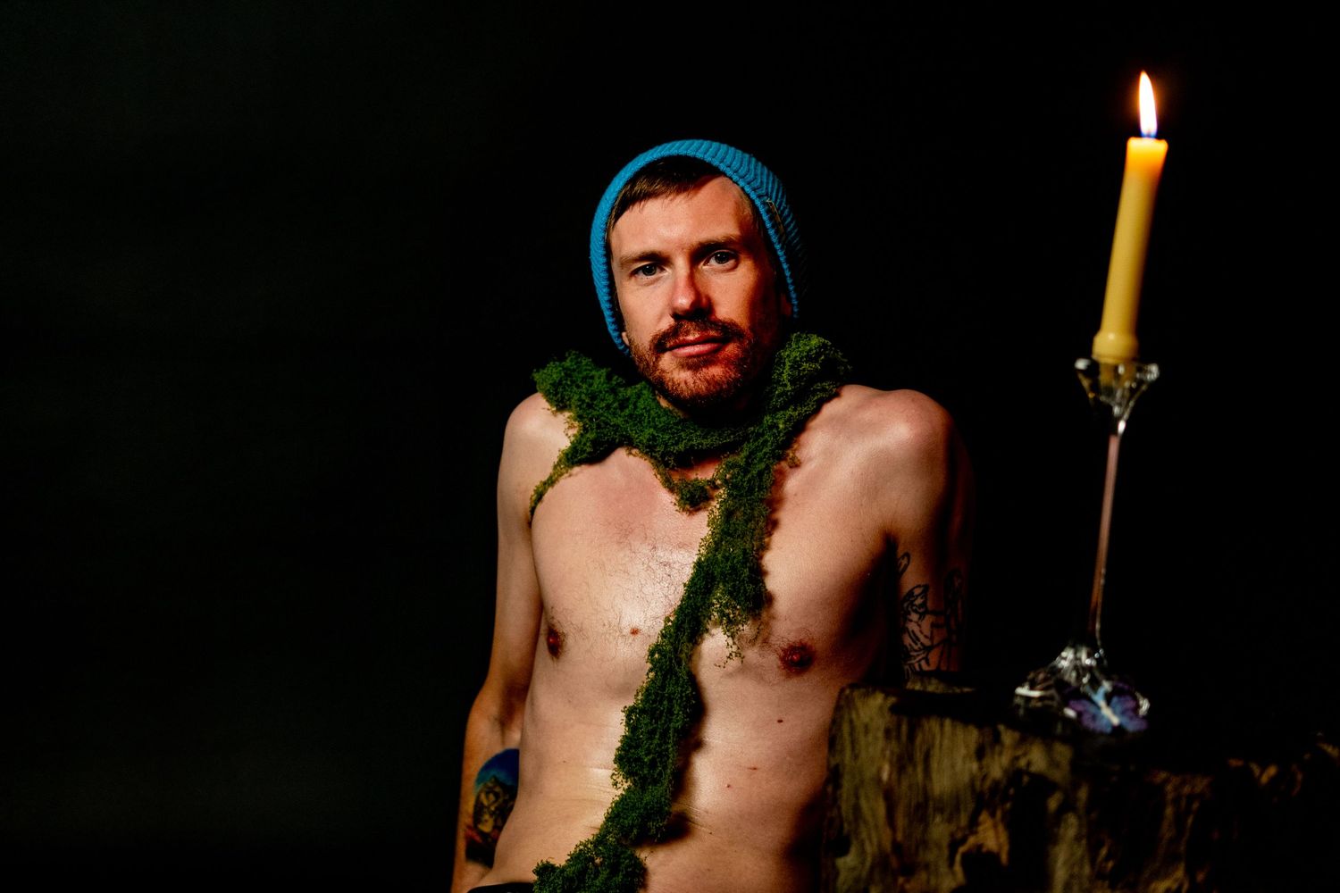 A shirtless person wearing a blue beanie and green garland poses in dramatic lighting next to a burning candle.