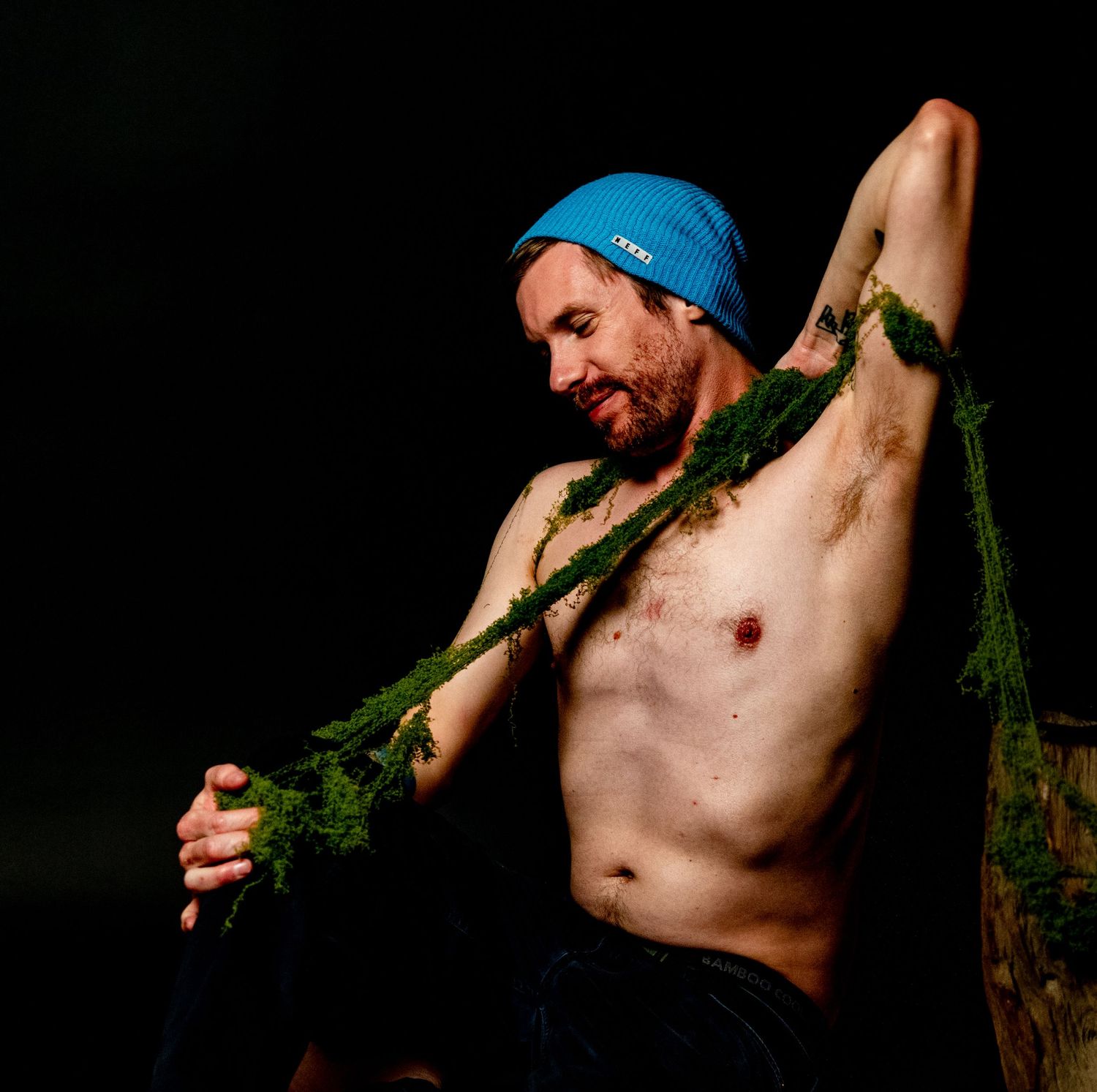 Shirtless person in dim lighting holding and posing with fresh green carrot tops against dark background.