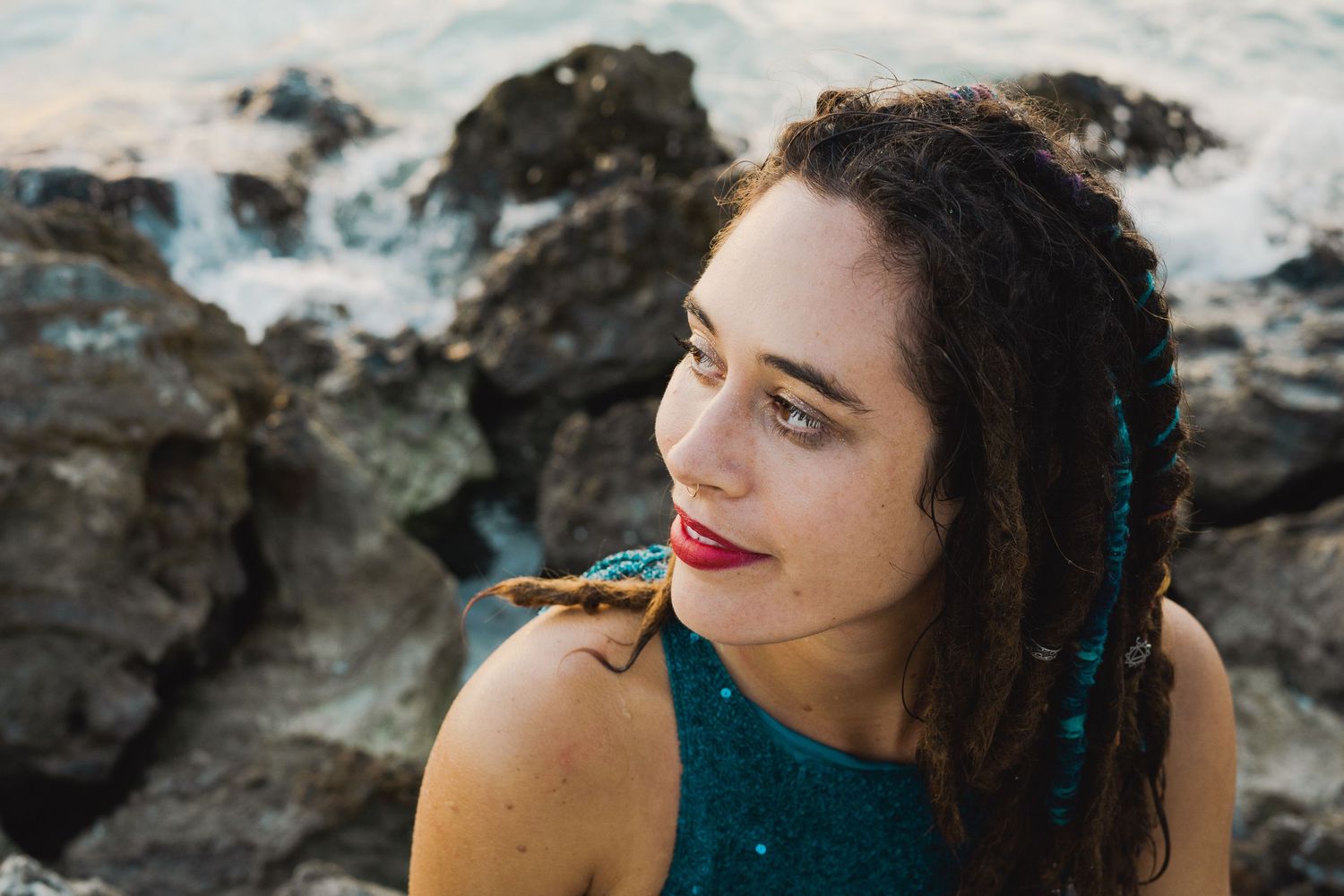 A person with long dark hair and red lipstick poses against a rocky outdoor background.