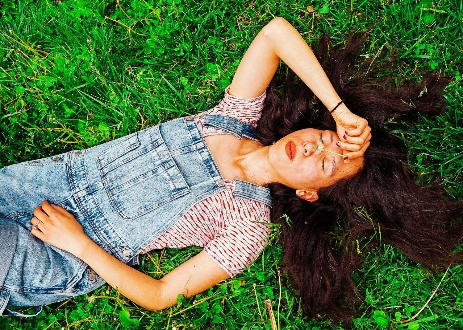 A photo of a woman with black hair sprawled out on the green grass. She is wearing a striped shirt with blue overalls.