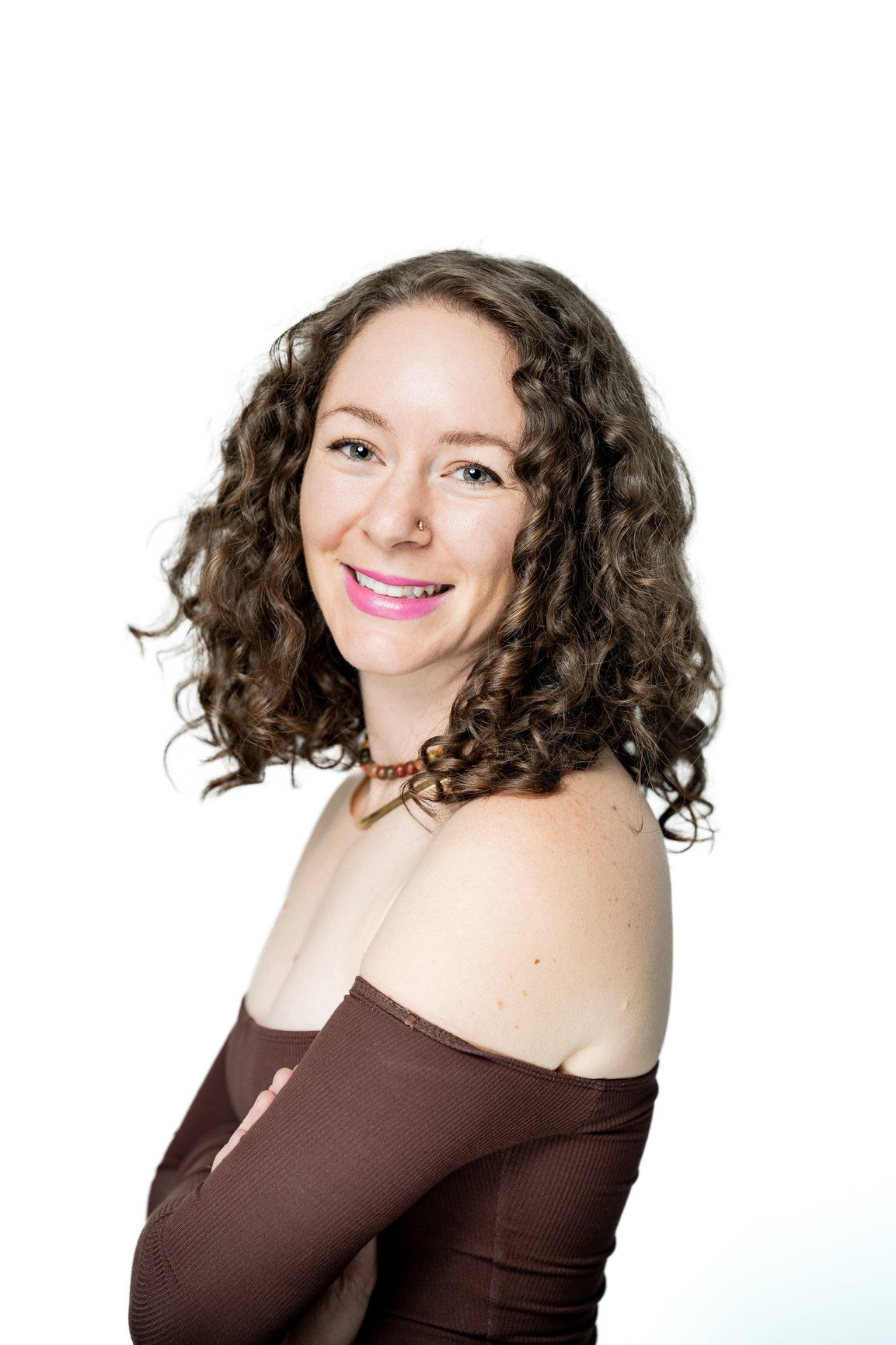 A photo of a woman with brown curly hair smiling and looking at the camera with a brown shirt draped off her shoulders.