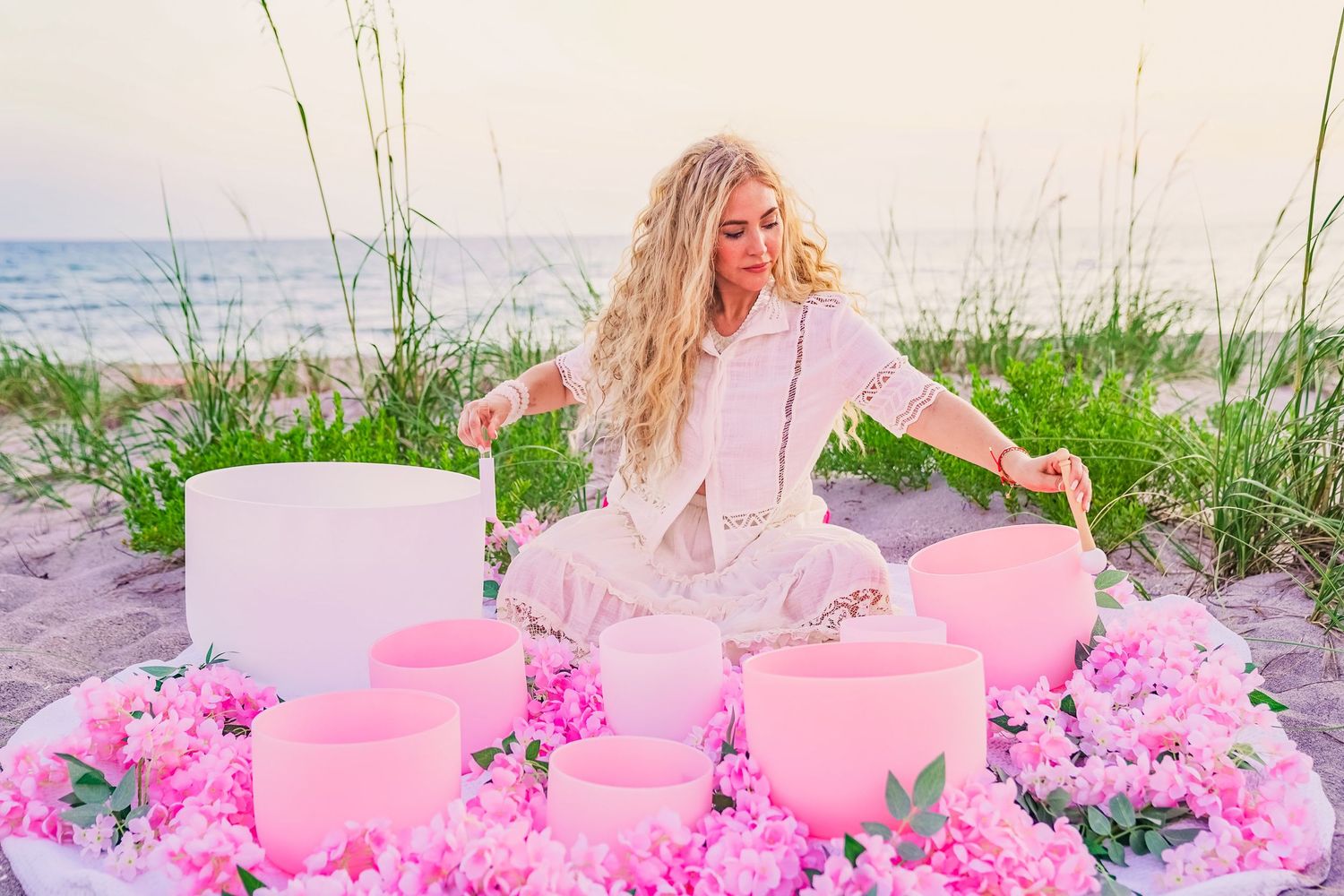 A photo of a blonde woman playing a white and pink singing bowls resting on pink flowers, she is on the beach and there is greenery.