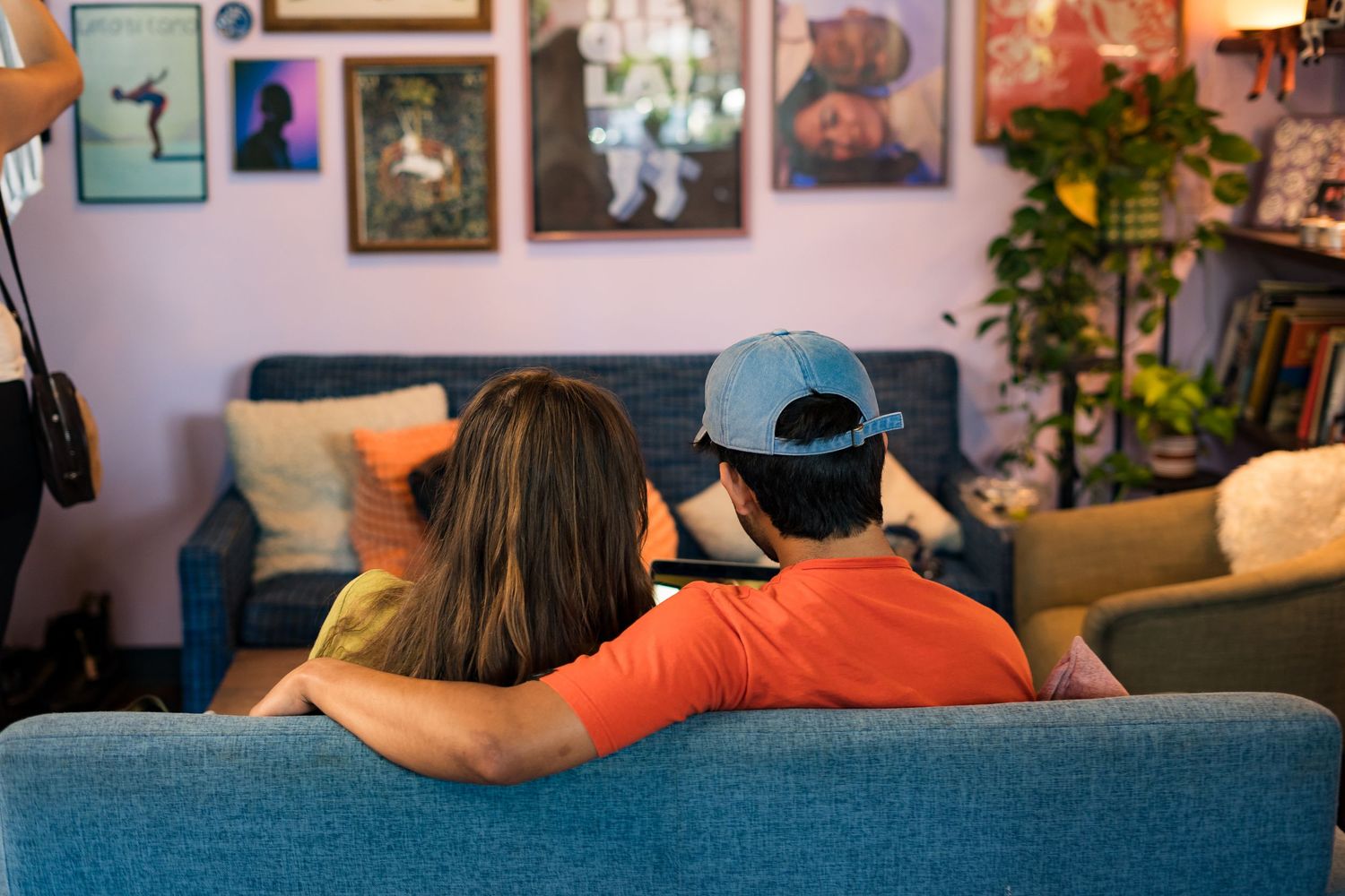 Couple sitting together on a blue sofa with arm around each other, viewed from behind in a cozy decorated living room.