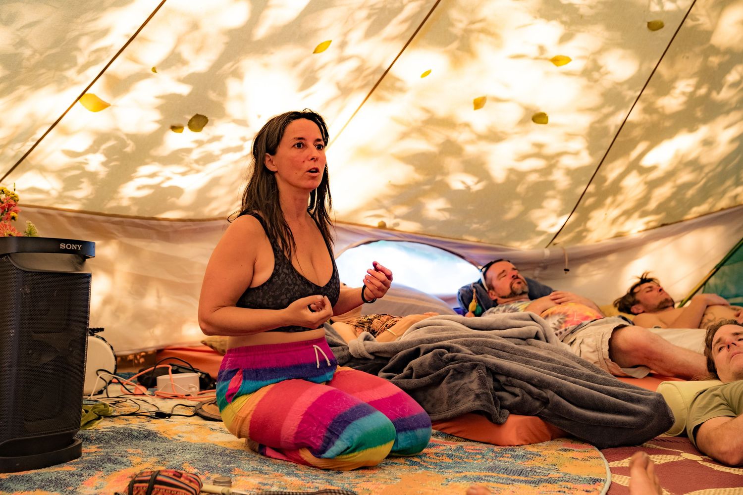 Woman meditating inside a glowing festival tent with colorful cushions while others rest in sleeping bags.