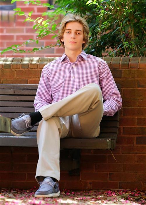 Young man in pink gingham shirt and khaki pants sitting relaxed on a bench outdoors near brick wall.