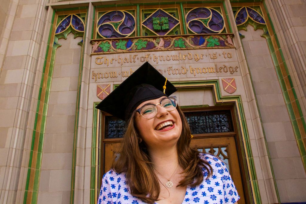 Smiling young woman in graduation cap poses in front of ornate university building entrance with stained glass.