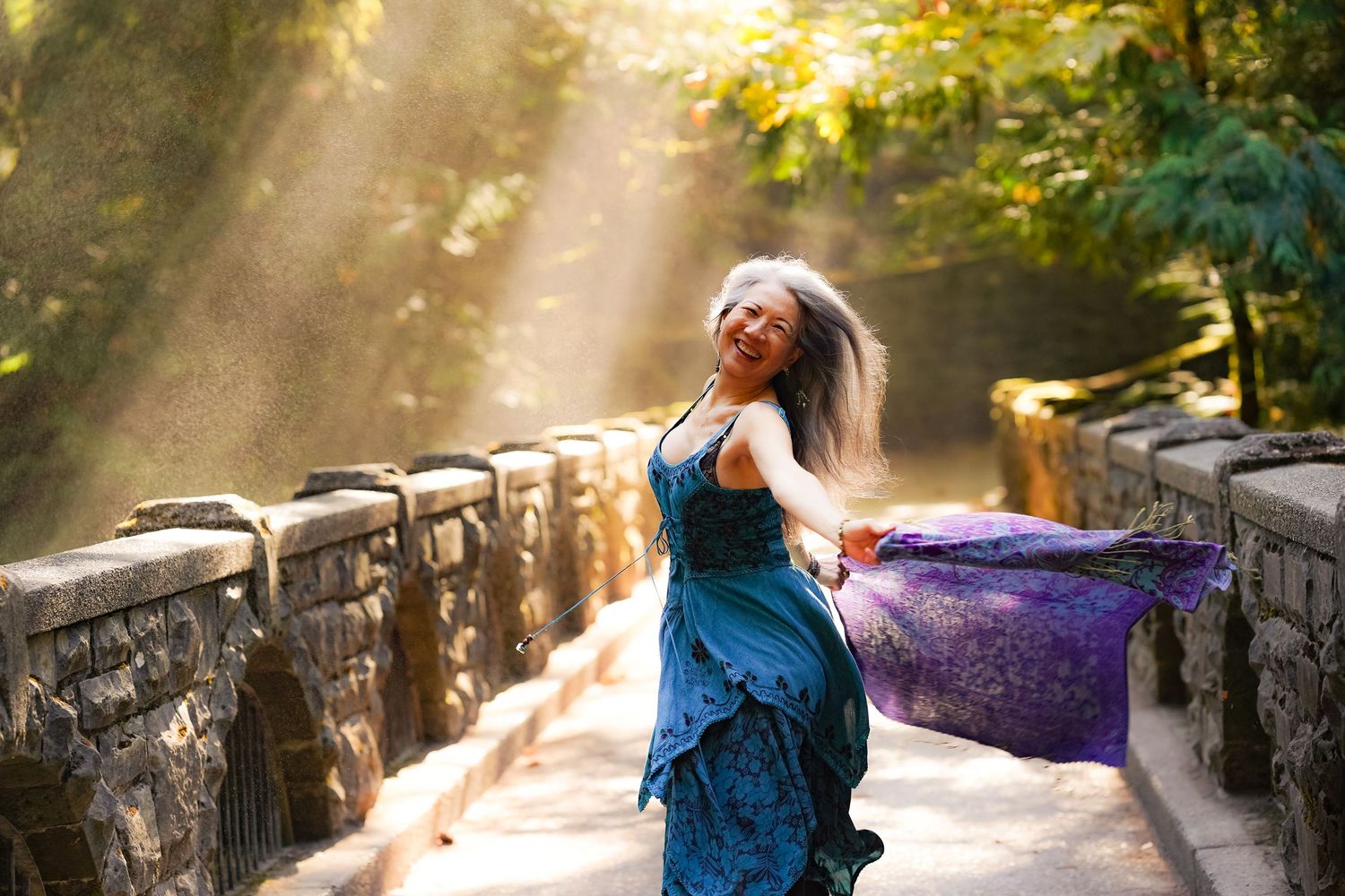 Woman in blue dress dancing on stone bridge with sunbeams filtering through trees in a magical outdoor setting.
