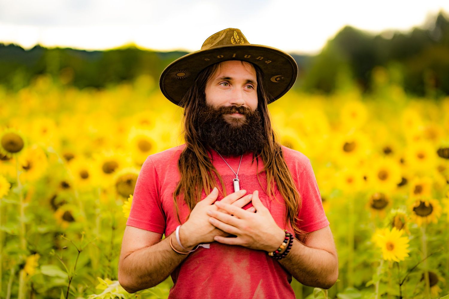Bearded man with long hair and wide-brim hat stands in a vibrant sunflower field, hands on chest.