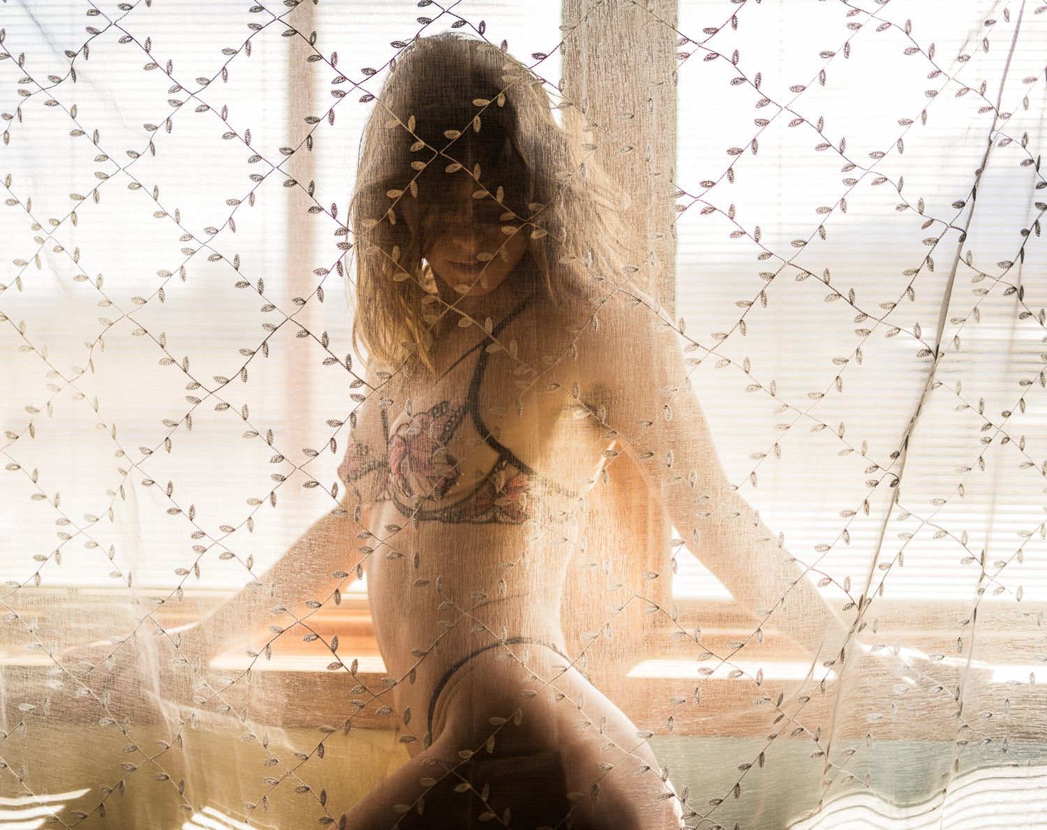 Woman in lingerie posing by a sunlit window with a floral curtain overlay creating a dreamy effect.