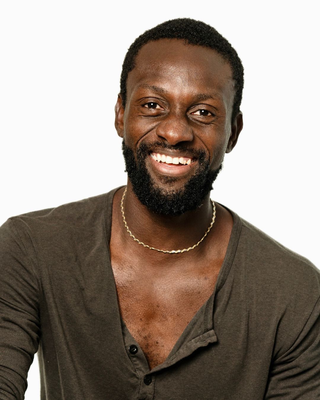 Smiling Black man with beard and gold necklace wearing an olive green V-neck shirt against white background.