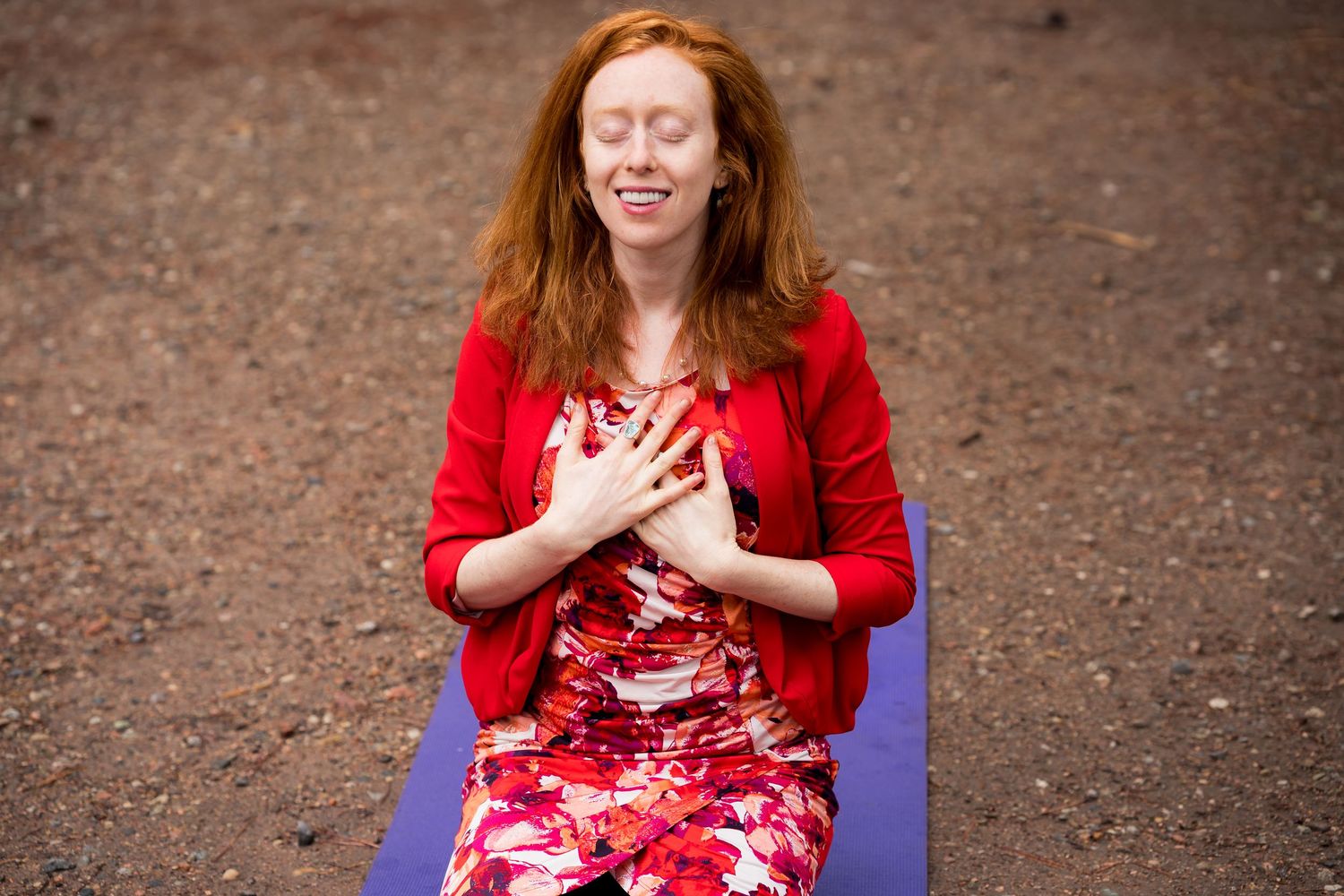 Red-haired woman in red cardigan sits on purple yoga mat outdoors, eyes closed, hands on heart in meditation pose.