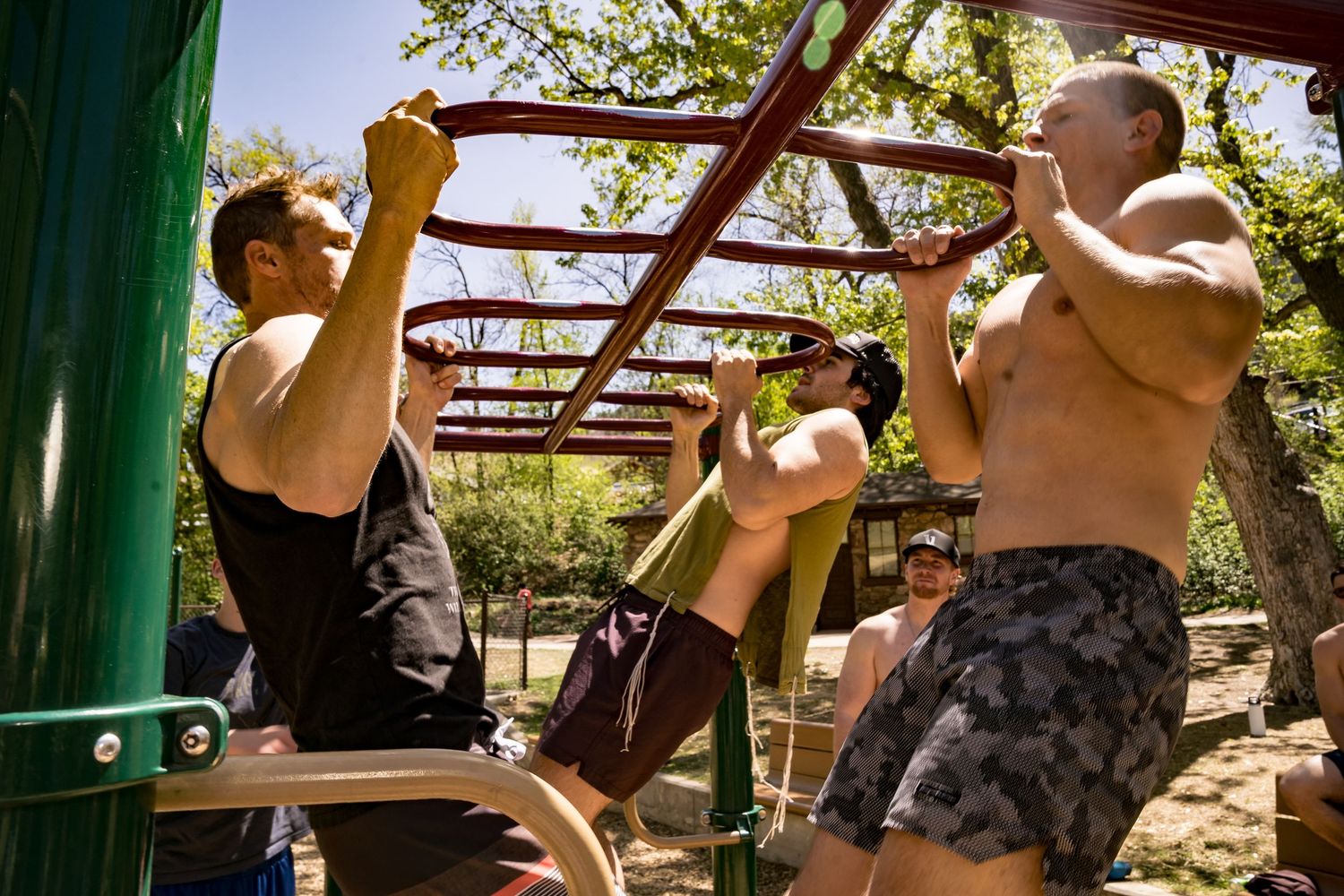 Men exercising on outdoor monkey bars in a sunny park workout area.