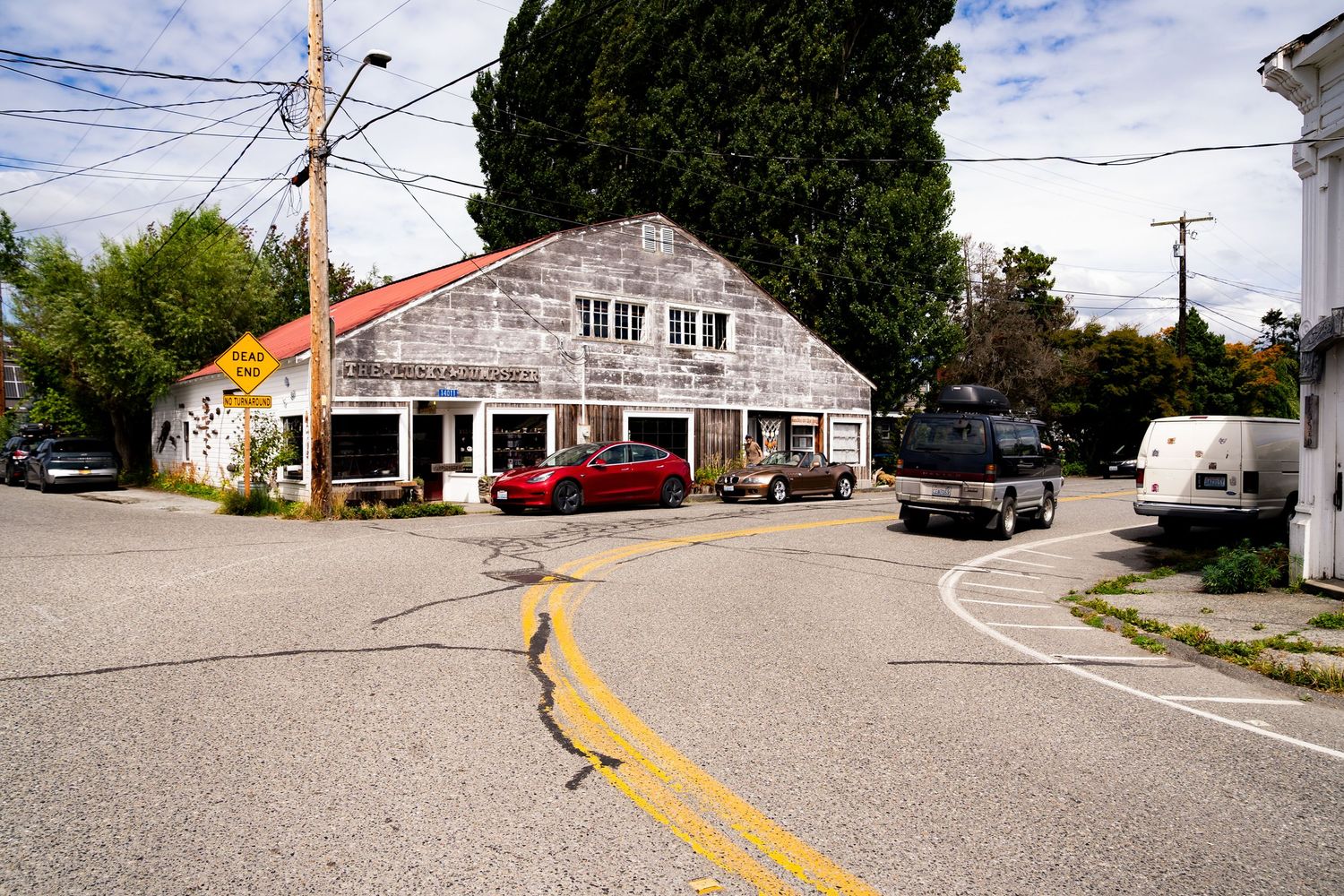 A weathered gray shingled building at a small-town intersection with parked cars and lush trees under a cloudy sky.
