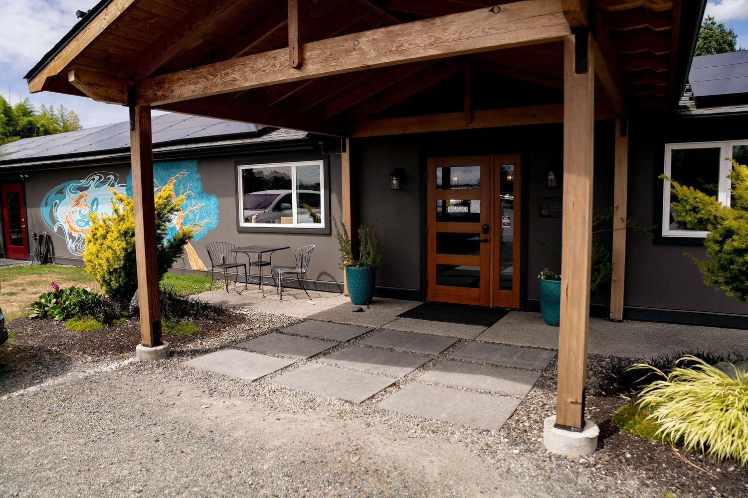 Modern building entrance with covered walkway, decorative mural, teal planters, and glass door on a gravel path.