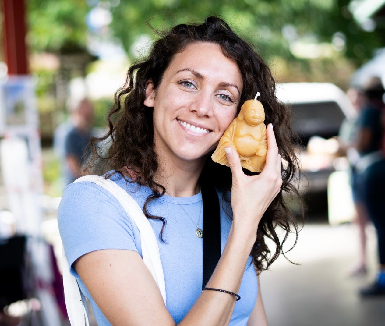 A photo of a pretty girl with wavy brown hair wearing a light blue shirt and a white bag holding her buddha candle up to her face
