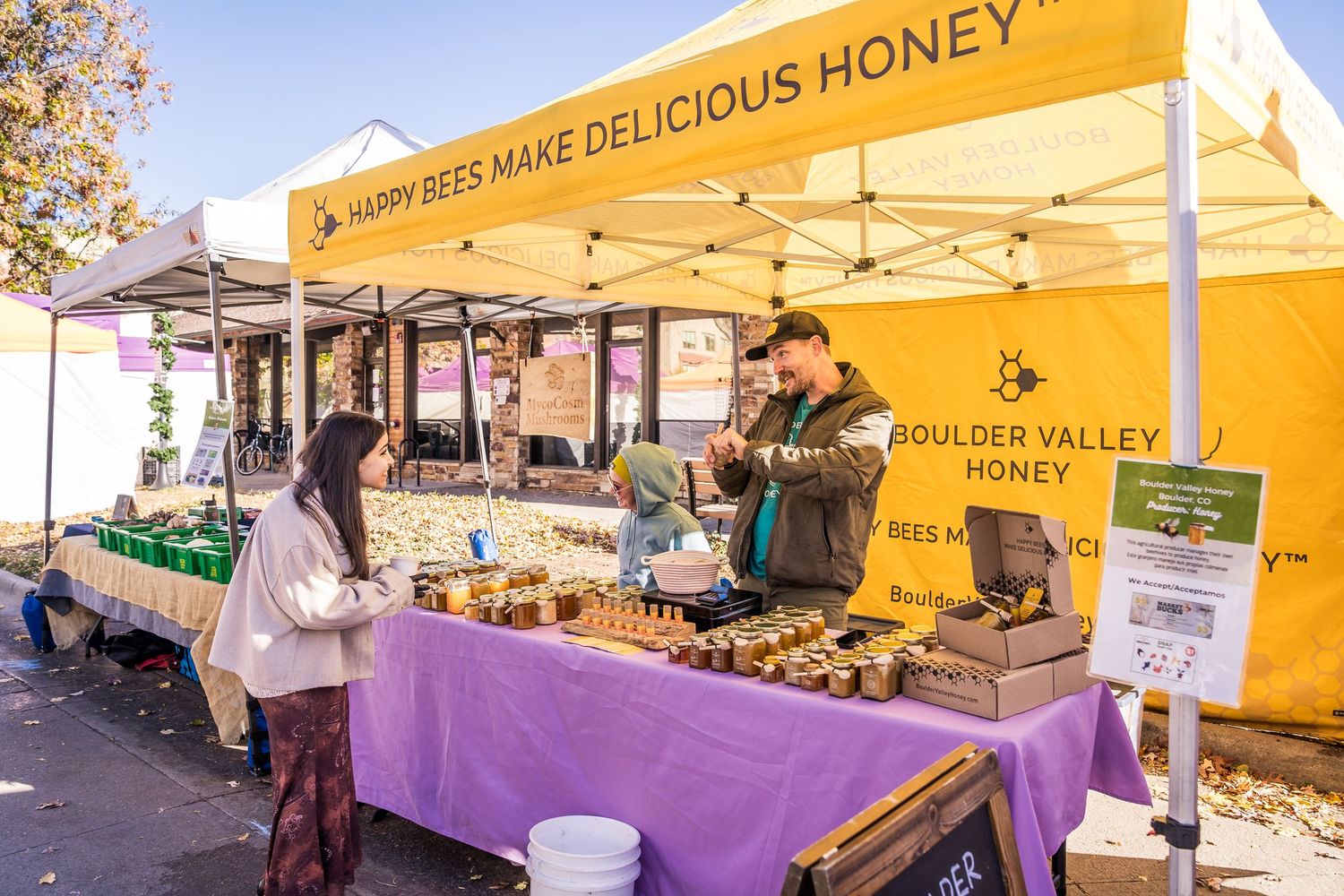 Happy Bees vendor handing a jar of local honey to an excited young girl at Boulder Farmers Market, showcasing Colorado-made honey