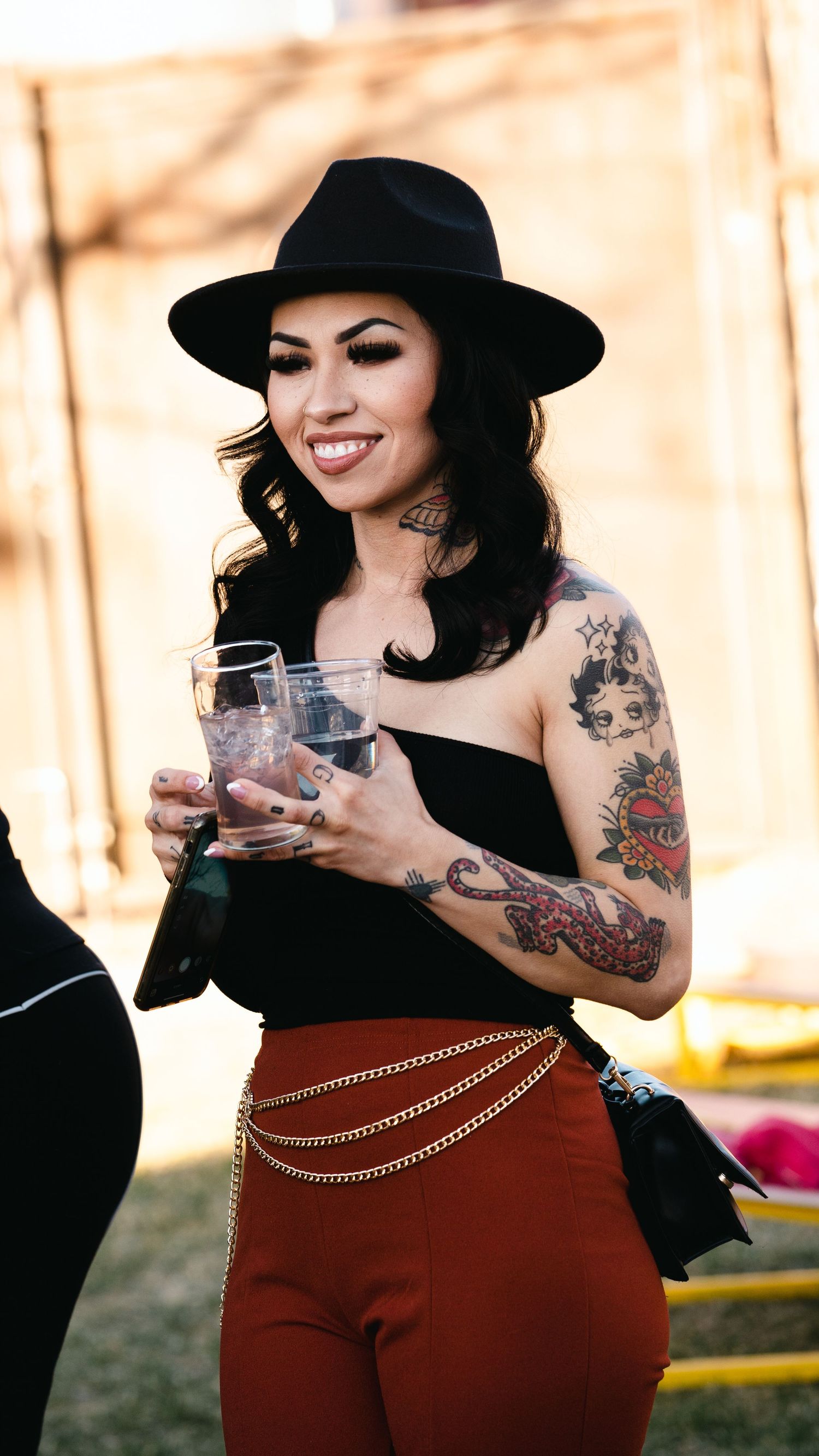 Smiling tattooed woman in black hat and strapless top holding a glass drink at an outdoor event.