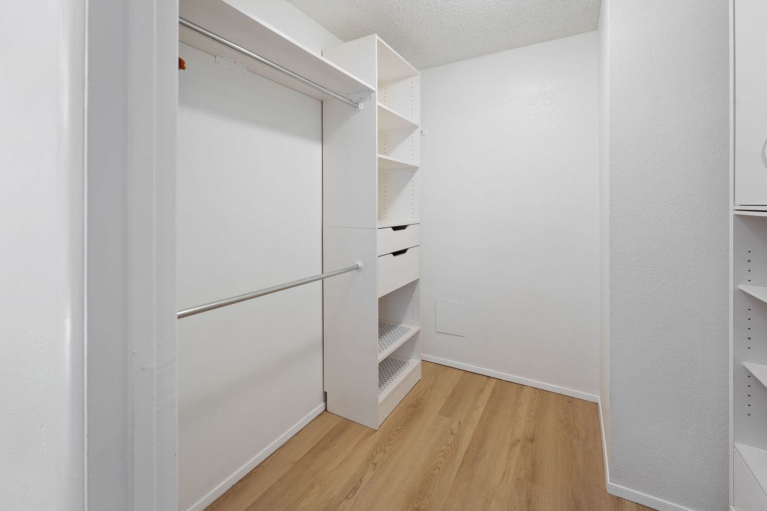 Spacious walk-in closet with white shelving, built-in drawers, hanging rods, and light hardwood flooring.